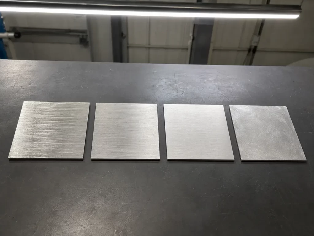 Four stainless steel sample panels showing different brushed finish textures — 100 grit coarse, 180 grit medium, 320 grit fine, and random non-directional grain, arranged side by side on a workbench.